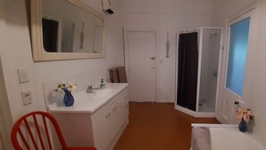 A narrow bathroom with a white sink, mirror, and shower. There is a red chair and a vase with flowers on the counter.