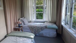 A bedroom with two beds, a window with curtains, a bedside table with a vase of flowers, and a framed picture on the wall.