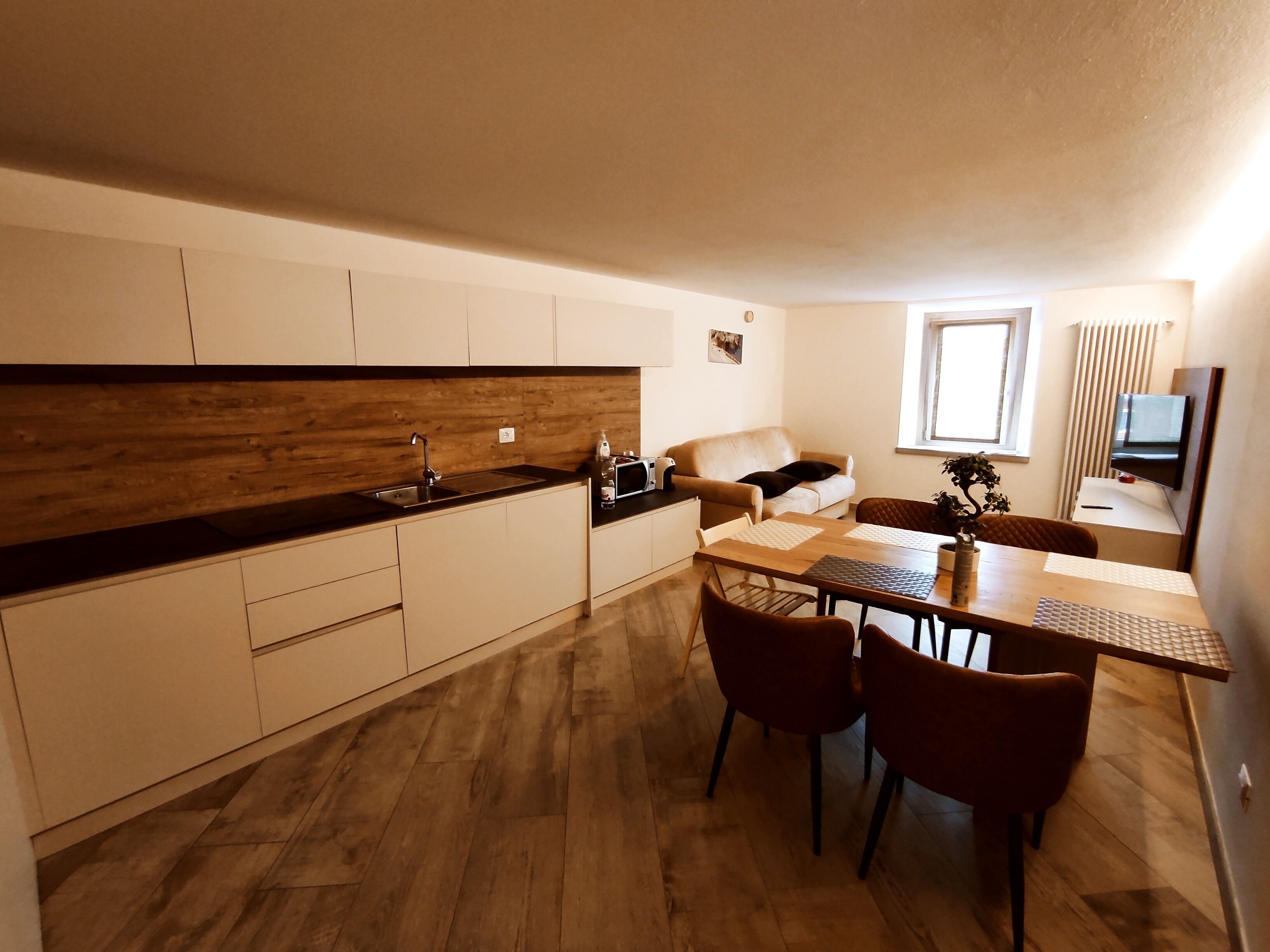 A modern kitchen with a wooden backsplash, a dining area with a table and chairs, a sofa, and a TV mounted on the wall.