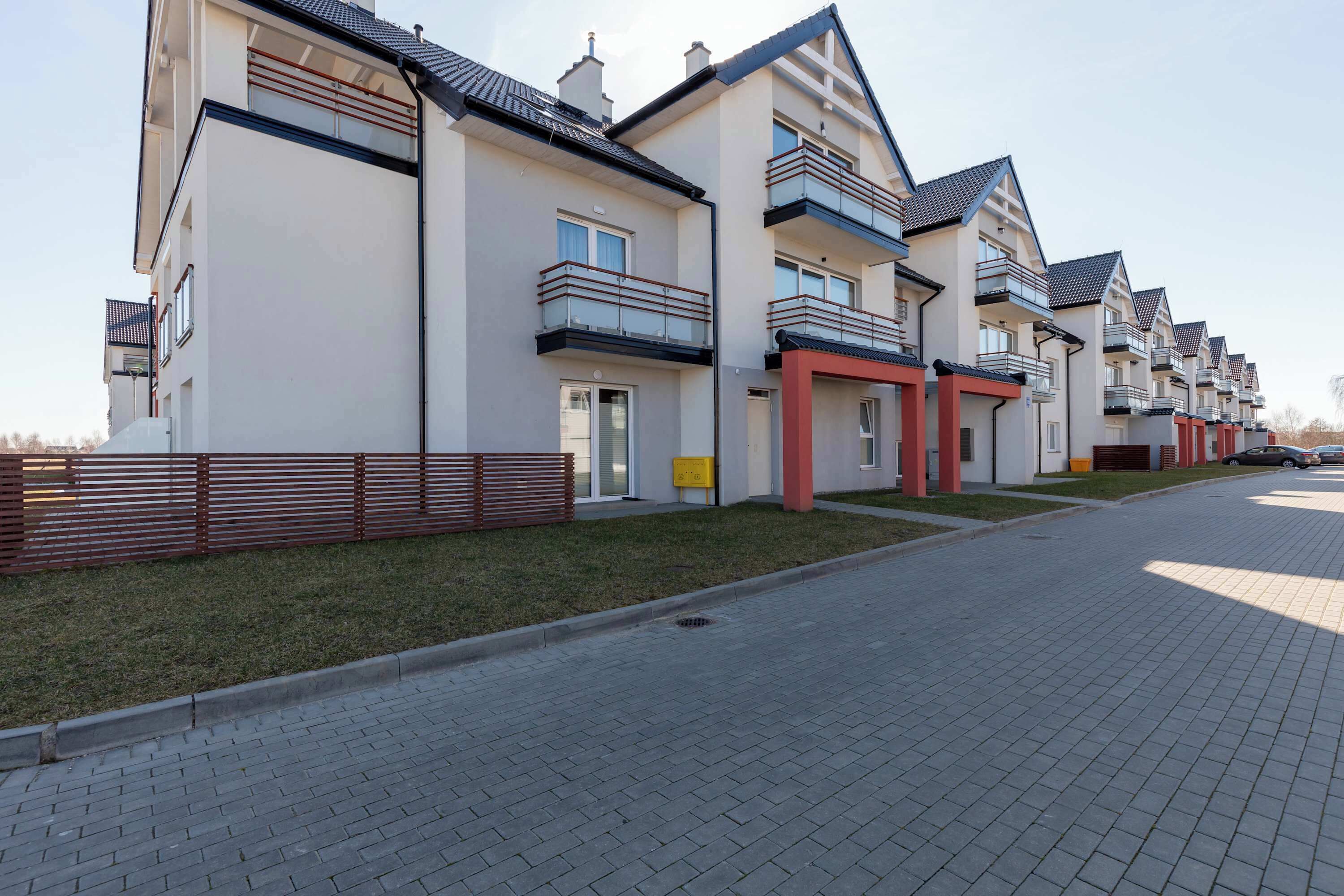 A row of modern apartment buildings with balconies, a paved driveway, and a wooden fence.