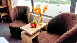 A room with two brown upholstered chairs, a small wooden table with a bottle of wine, a glass, and a plate of fruit. A window offers a view of a river with boats. - Pearl Beach Hotel Quy Nhon (Quy Nhon Dong)