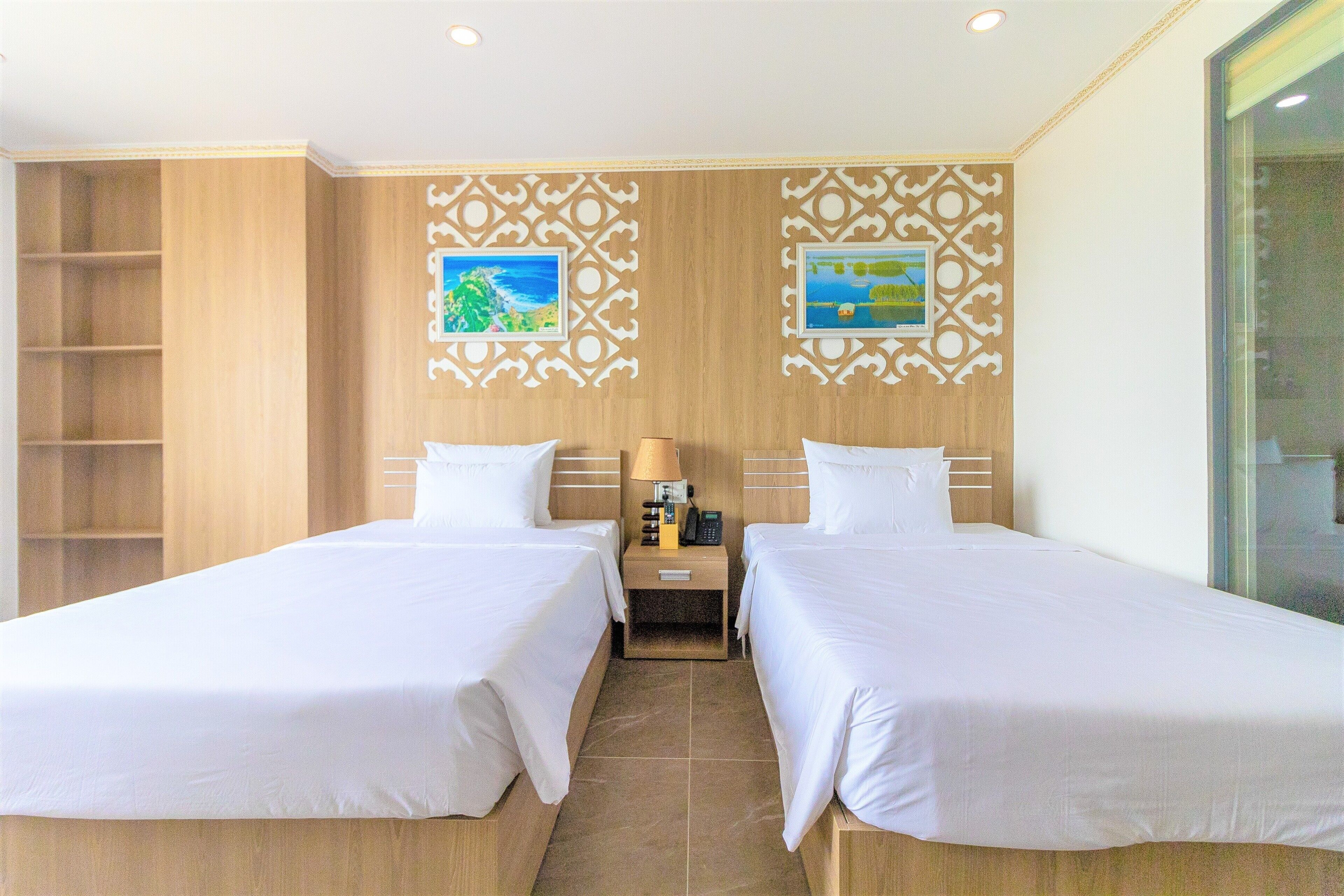 A hotel room with two single beds, a wooden wardrobe, a small table with a lamp, and decorative wall panels.