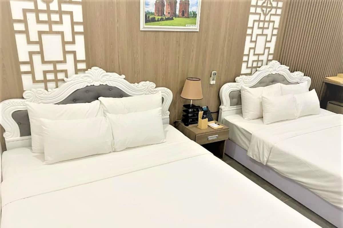 Una habitación de hotel con dos camas, cada una con ropa blanca y cabeceros decorativos. Hay una mesita de noche con una lámpara y un teléfono entre las camas.