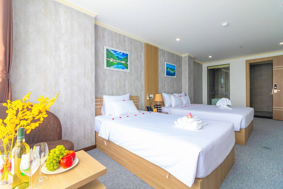 Habitación de hotel con dos camas, un escritorio de madera, un florero con flores, un plato de fruta y dos cuadros en la pared.