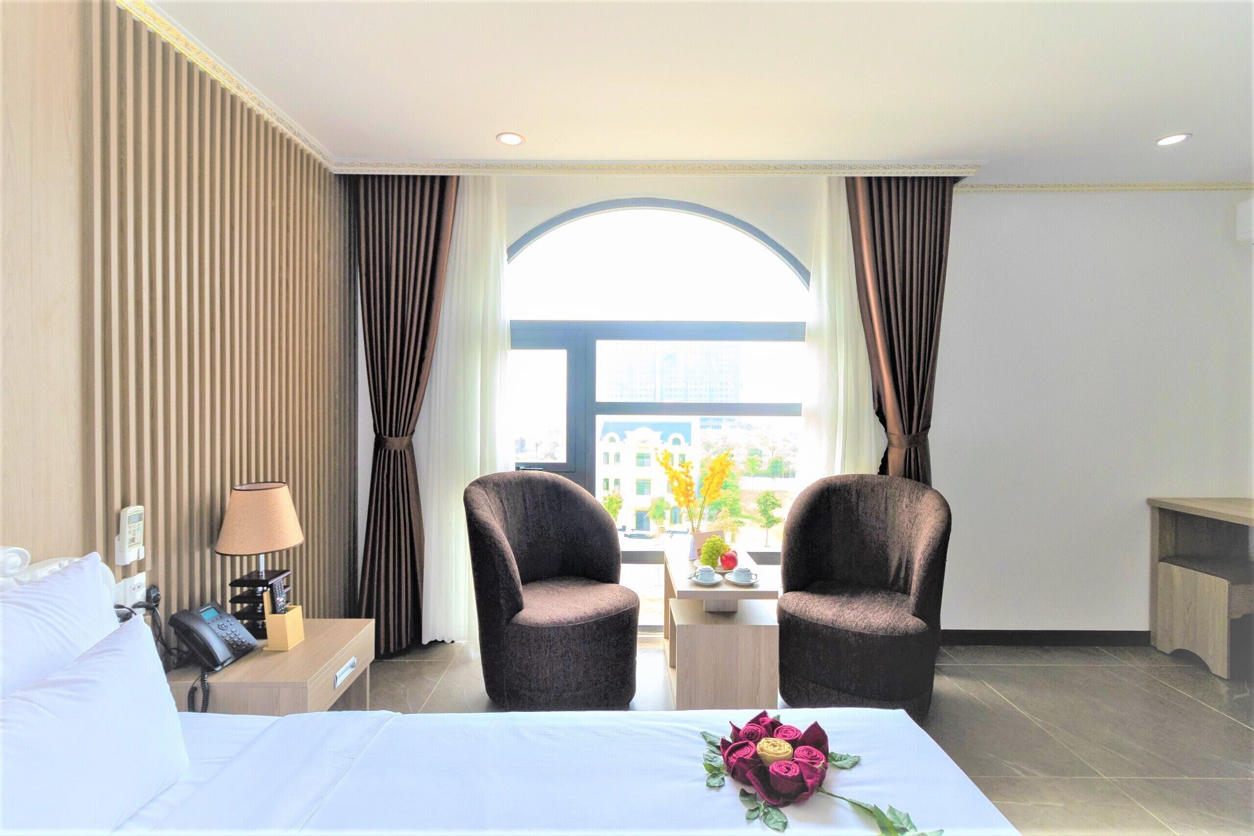 A hotel room with a bed, two armchairs, a small table with a vase of flowers, a lamp, and a view of the city through a large window.