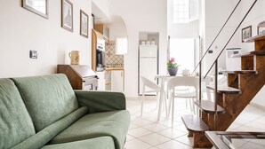 A modern living room with a green sofa, a dining area with white chairs, a kitchen with white cabinets, and a staircase with wooden steps.
