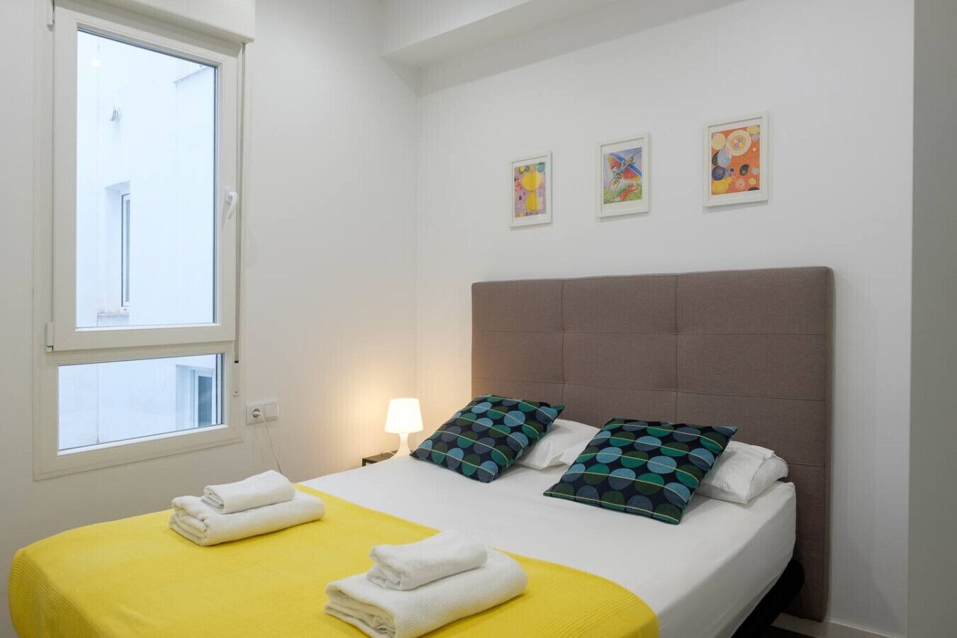 Une chambre d’hôtel avec un lit, deux oreillers, une lampe de chevet et trois œuvres d’art encadrées accrochées au mur.