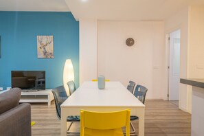 A dining area with a white table, blue chairs, a yellow chair, a clock, and a TV on a stand.