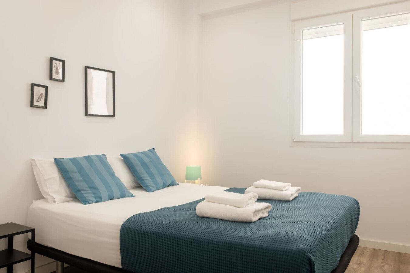 A neatly made bed with blue and white bedding, a small bedside table with a lamp, and a window with white frames.