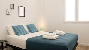 A neatly made bed with blue and white bedding, a small bedside table with a lamp, and a window with white frames. - Bright, Clean & Very Comfortable Apartment (Valencia)
