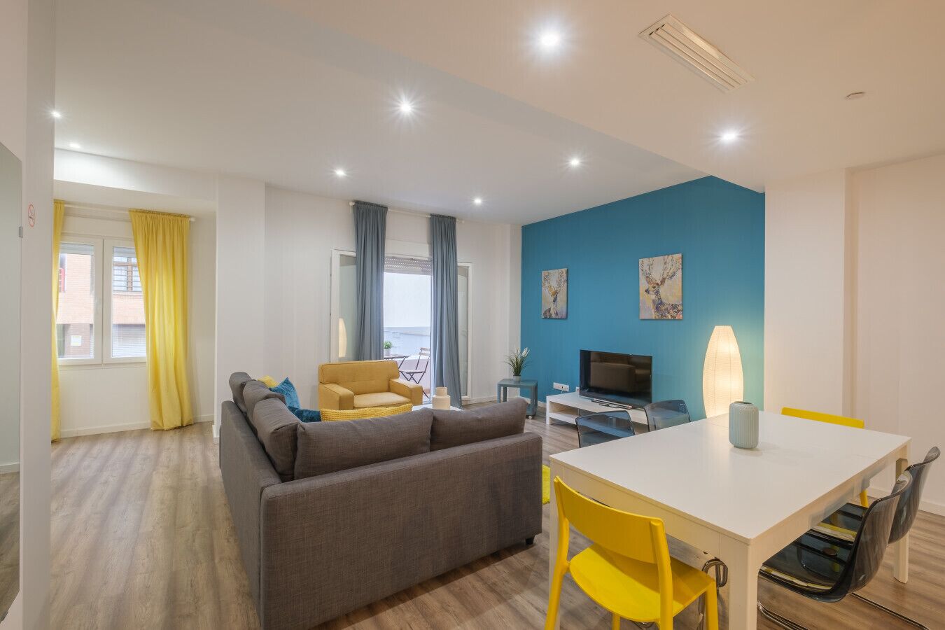 A modern living room with a sofa, a yellow armchair, a dining table with chairs, a TV, and artwork on the wall.
