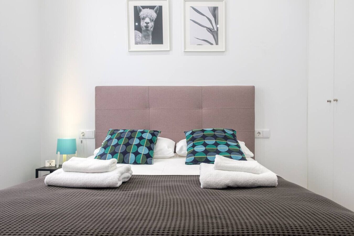 Een net opgemaakt bed met twee kussens, een bedlamp en een ingelijst foto aan de muur.