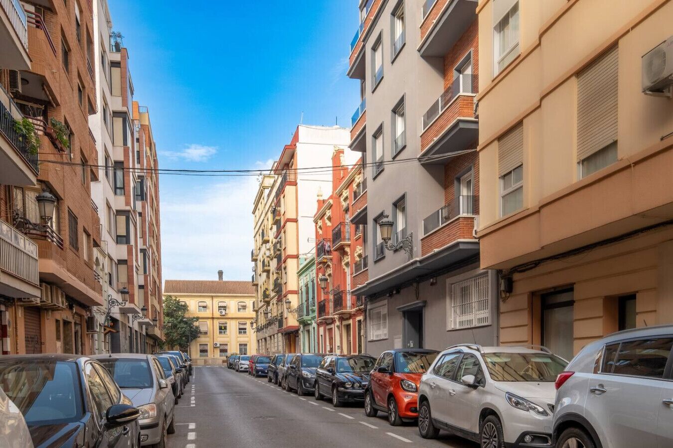 Eine Straße gesäumt von mehrstöckigen Gebäuden, parkenden Autos und einem strahlend blauen Himmel.