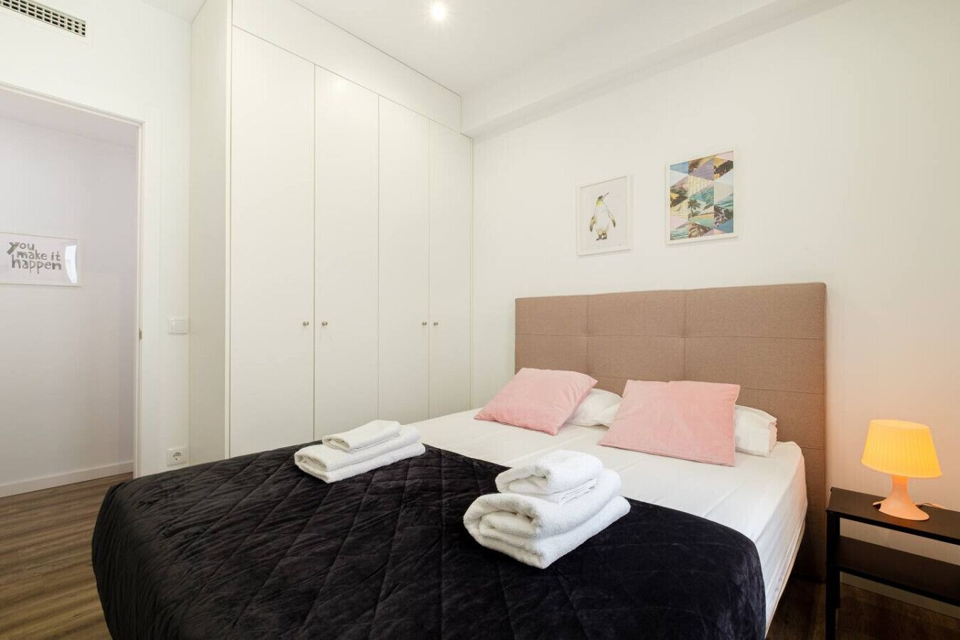 Een hotelkamer met een bed, twee kussens, een nachtkastje met een lamp en een garderobe.