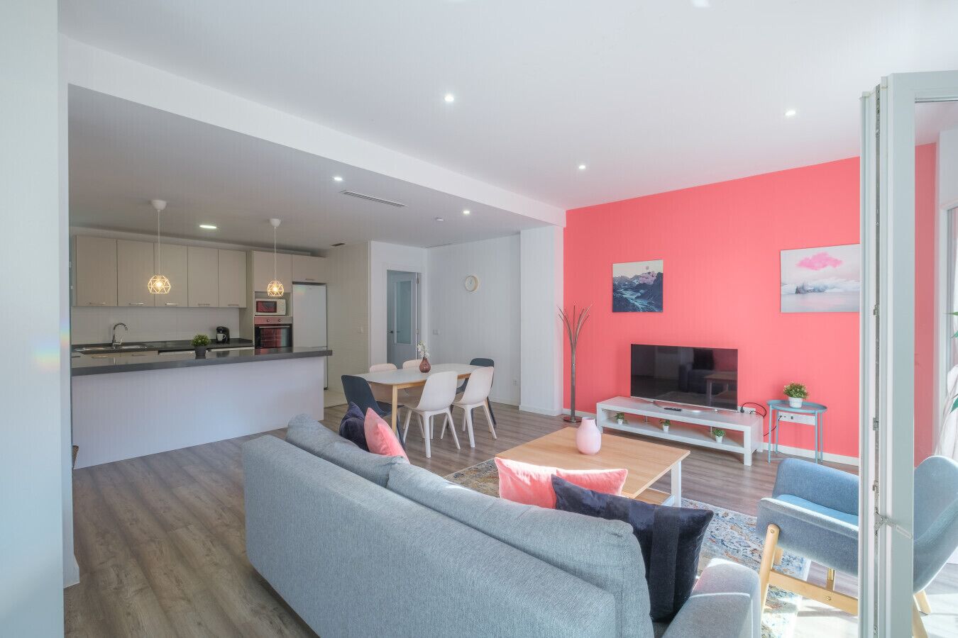 A modern living space with a grey sofa, a dining area, a TV, and a bright pink wall.
