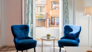 Een kamer met twee blauwe fauteuils, een kleine ronde tafel met een plant, en uitzicht op een balkon met een leuning en plantenbakken.