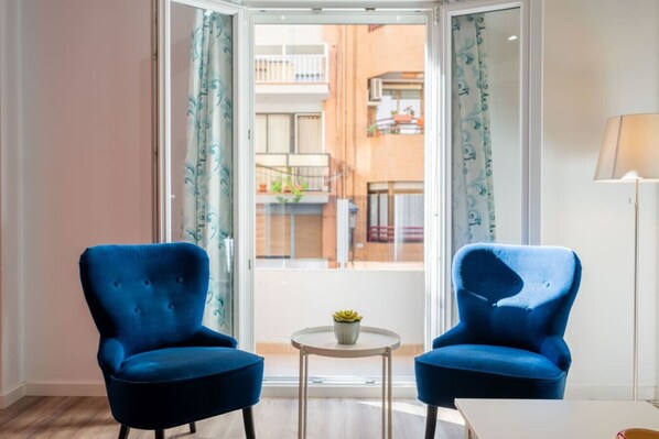 A room with two blue armchairs, a small round table with a plant, and a view of a balcony with a railing and potted plants.