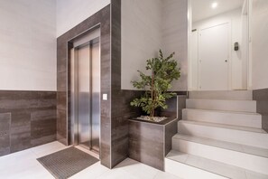 Un ascensor moderno con acabado metálico, una escalera de peldaños blancos y una jardinera con follaje verde.
