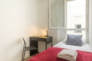 Une petite pièce, bien rangée, avec un lit, un bureau et une chaise. Une fenêtre laisse entrer la lumière naturelle, et une serviette rouge est posée sur le lit.