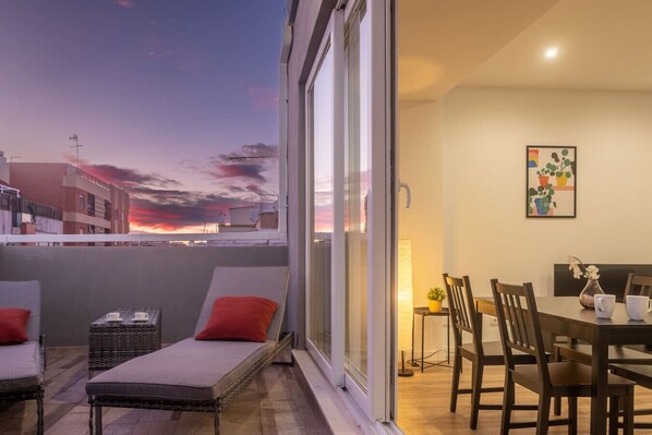 Un balcon avec vue sur le coucher du soleil, un coin repas avec des chaises et une table, et un lit avec des oreillers.