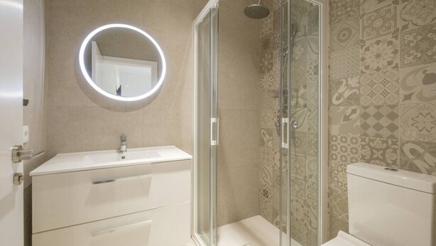 A modern bathroom with a glass-enclosed shower, a round mirror with a light, and a patterned tile wall. - ≬ Clean & Cozy Room close to City Centre ≬ (Valencia)