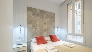 A bedroom with a bed, two red pillows, a beige quilt, a bedside table, a nightstand, and a patterned wall. - ≬ Clean & Cozy Room close to City Centre ≬ (Valencia)