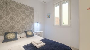 Um quarto com papel de parede estampado, uma cama com duas almofadas, uma mesinha de cabeceira, uma janela e um quadro na parede.