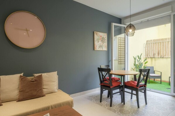 A modern living room with a sofa, a dining table with chairs, a large round clock, and a view of a garden through a glass door.