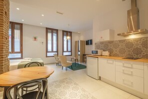 Una cocina moderna con encimera de madera, armarios blancos y campana extractora de acero inoxidable. Hay una zona de comedor con mesa y sillas de madera, y una pequeña isla de cocina con fregadero.