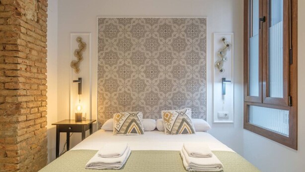 Une chambre d’hôtel avec un mur en briques, une tête de lit à motifs, un lit avec une literie blanche et des coussins décoratifs, une table de chevet avec une lampe et une fenêtre avec des stores.