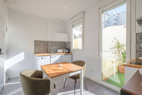 A compact kitchen with a wooden table and two chairs, a window with a view of buildings, and a balcony with a potted plant.