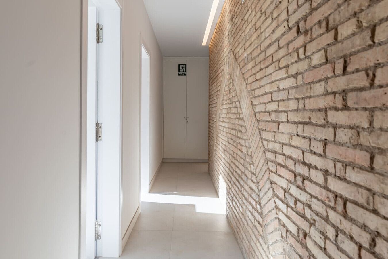 Un couloir étroit, avec un mur de briques d’un côté et des portes blanches de l’autre.