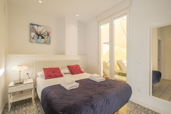 A hotel room with a large bed, a nightstand with a lamp, a painting on the wall, and a sliding door leading to a balcony with chairs.