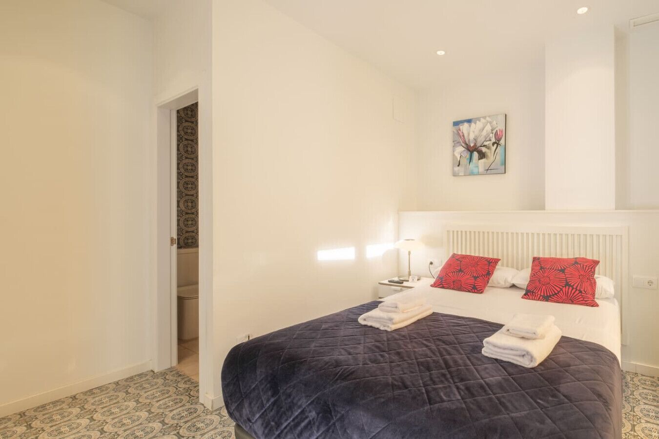 Una cama bien hecha con colcha oscura, almohadas blancas y una almohada roja con estampado floral sobre un cabecero blanco.