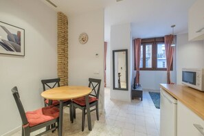 A compact living space with a dining area featuring a wooden table and black chairs, a kitchen with a microwave, and a brick accent wall.