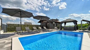Une piscine entourée d’une haie et agrémentée de chaises longues et de parasols, avec une cabane en bois à proximité.