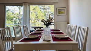 A dining area with a table set for four, chairs, a vase with flowers, and a view of trees through large windows. - MILN - Apartment in holiday rental, beach center of Carnac - T11 (Carnac)