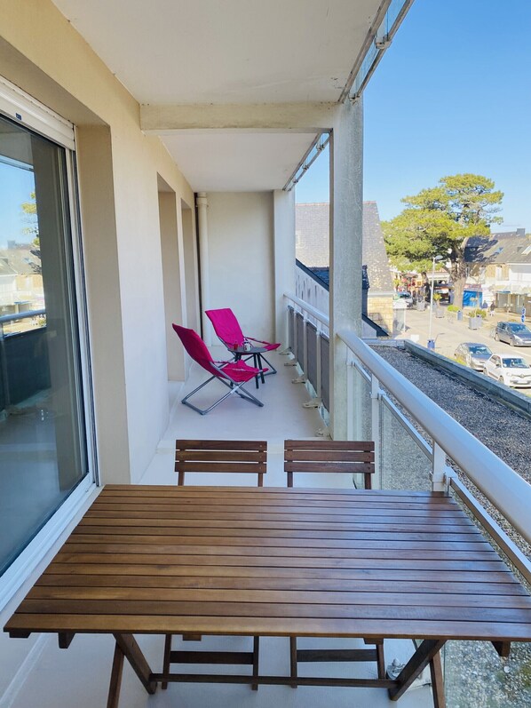 A balcony with a wooden table and two red chairs, overlooking a street with parked cars and buildings.