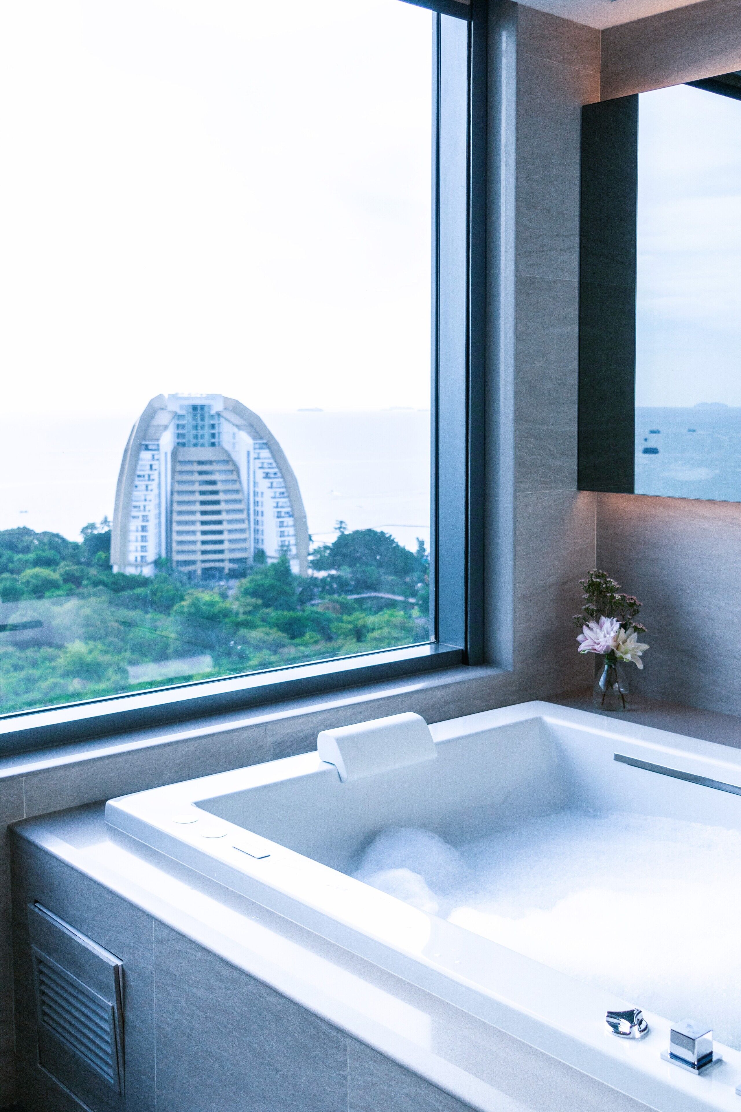 A modern bathroom with a large bathtub, a window with a view of a building, and a vase with flowers.