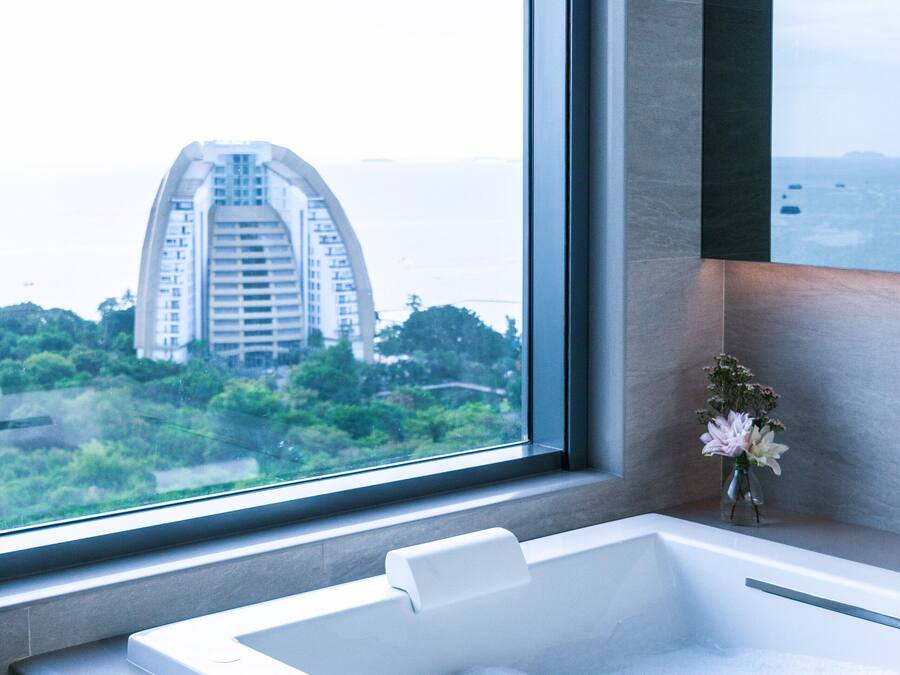 A modern bathroom with a large bathtub, a window with a view of a building, and a vase with flowers.