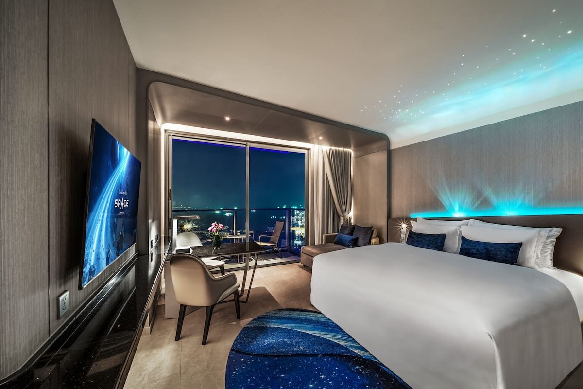 A modern hotel room with a large bed, a desk, and a view of the city at night.