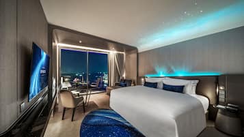 A modern hotel room with a large bed, a desk, and a view of the city at night.