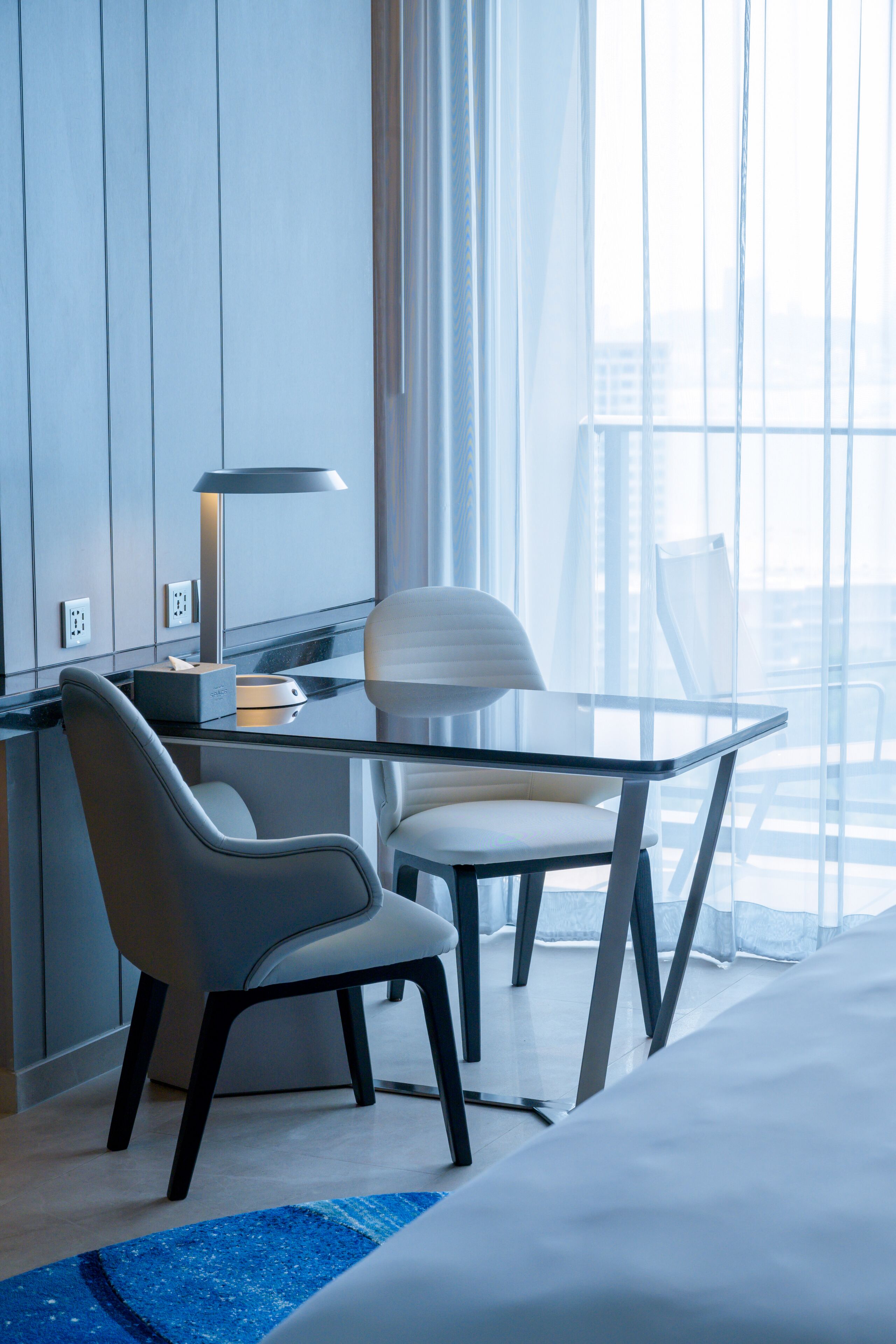 A modern hotel room with a glass table, two chairs, a desk with a lamp, and a large window offering a city view.