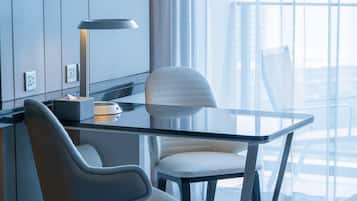 Une chambre d’hôtel moderne avec une table en verre, deux chaises, un bureau avec une lampe et une grande fenêtre donnant sur la ville.