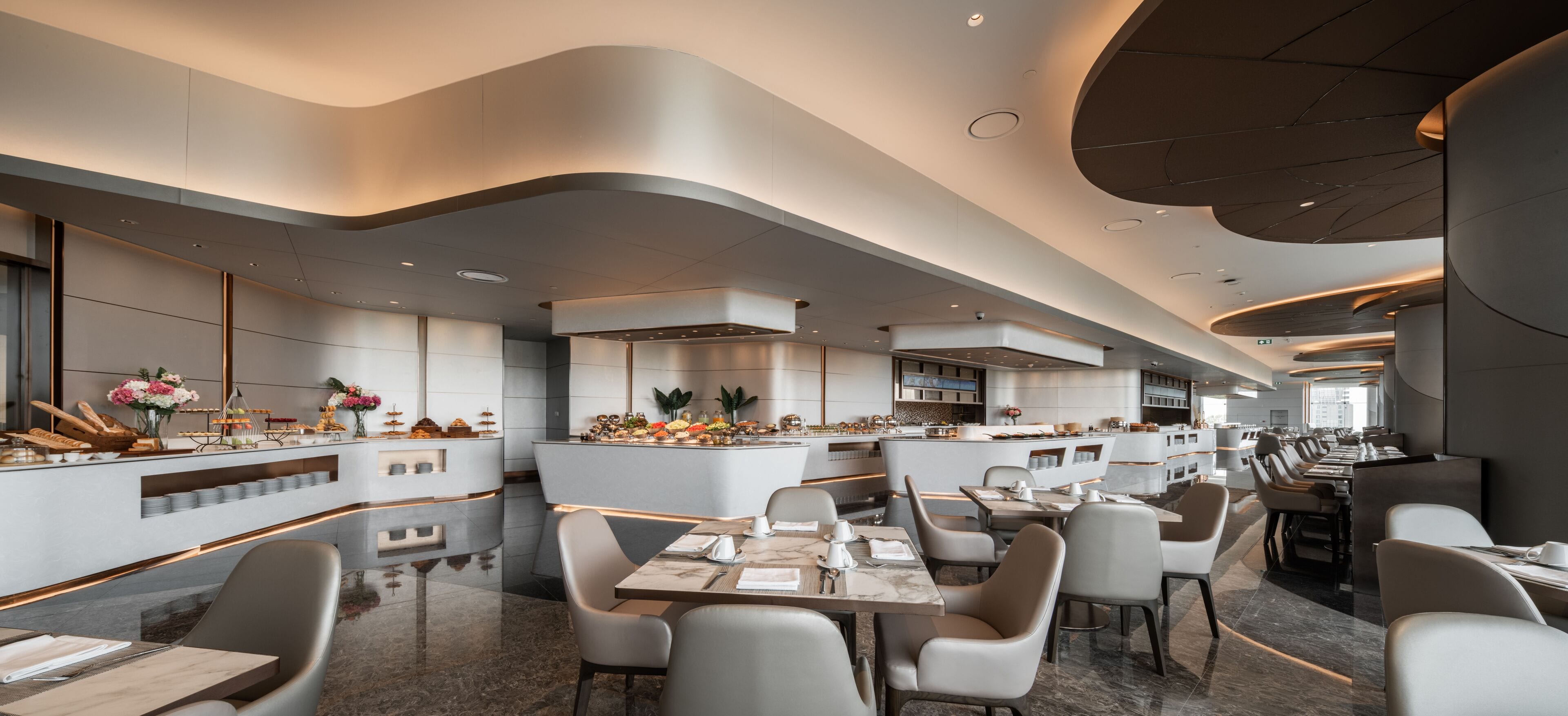 A modern restaurant with a sleek, curved ceiling design and a long dining area with multiple tables and chairs.