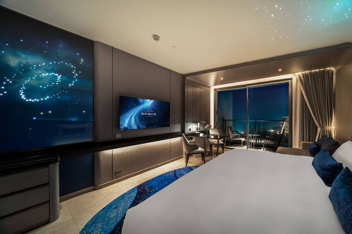 A modern hotel room with a large bed, a desk, and a TV displaying a space-themed wallpaper.