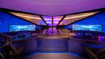 A gaming room with two large monitors, a steering wheel, and a racing game displayed on the screens.
