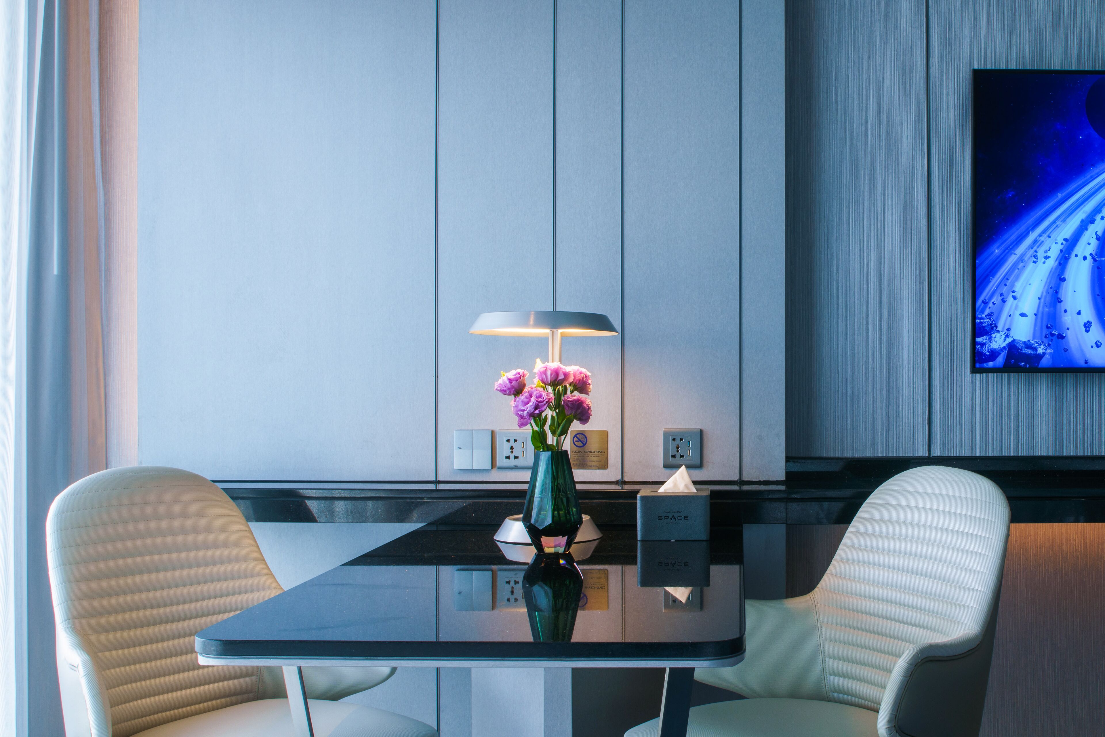 Una habitación de hotel moderna con una mesa de vidrio, dos sillas blancas, un florero verde con flores, una lámpara y un televisor en la pared.