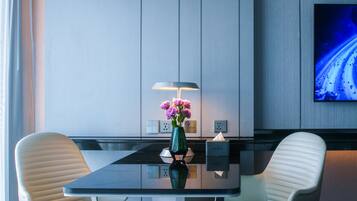 Una habitación de hotel moderna con una mesa de vidrio, dos sillas blancas, un florero verde con flores, una lámpara y un televisor en la pared.