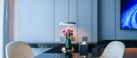 A modern hotel room with a glass table, two white chairs, a green vase with flowers, a lamp, and a TV on the wall.